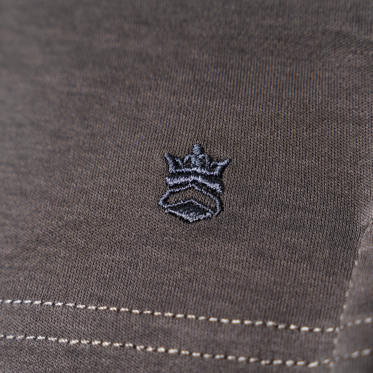 Close-up of a dark gray textured fabric with a small embroidered crown logo in black and gray, plus white contrast stitching near the bottom. Thomas Maine T-Shirts available at Sakks Menswear in Southampton.