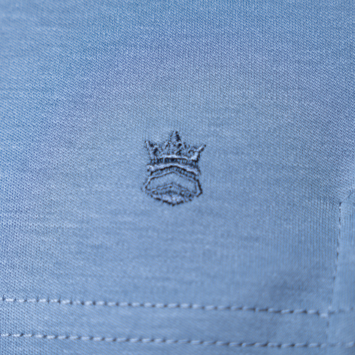 Close-up of light blue fabric with a small embroidered crown logo in a darker blue-gray thread. Thomas Maine T-Shirts available at Sakks Menswear in Southampton.