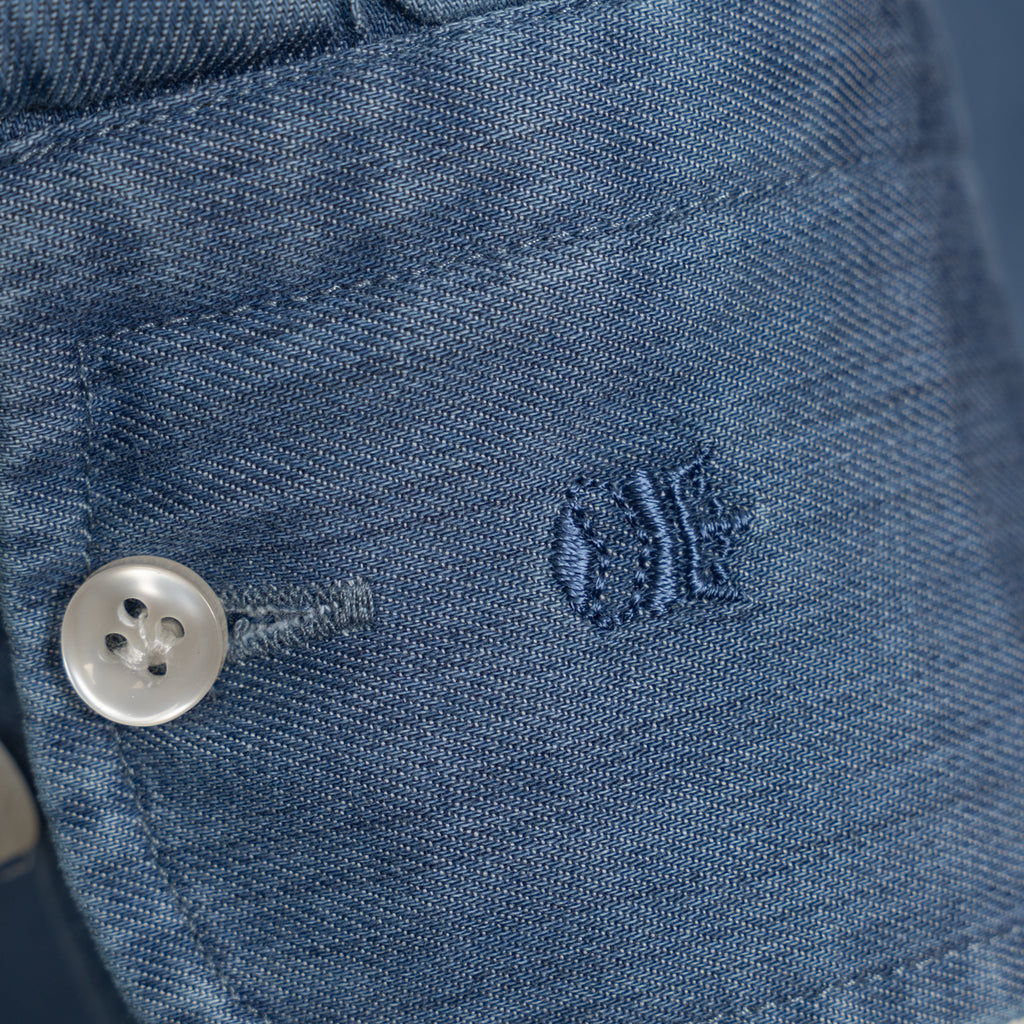Close-up of a blue denim garment showing a silver button, stitching, and a small embroidered logo on the fabric. Thomas Maine Shirt available at Sakks Menswear in Southampton.