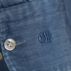 Close-up of a blue denim garment showing a silver button, stitching, and a small embroidered logo on the fabric. Thomas Maine Shirt available at Sakks Menswear in Southampton.