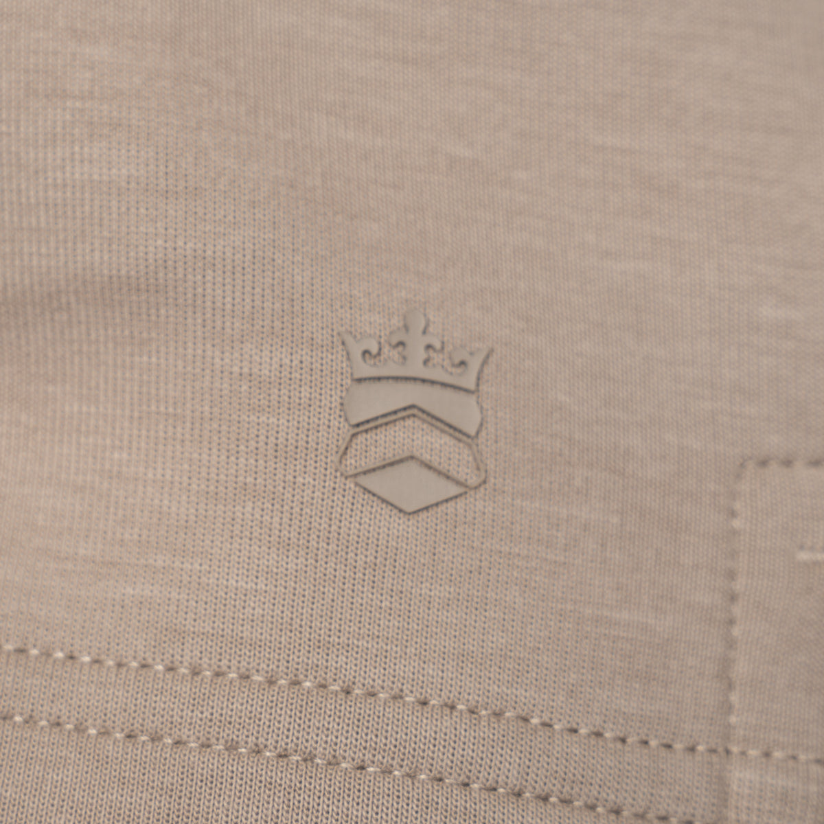 Close-up of a light beige knit fabric with a tonal embroidered crown logo centered on the chest. Thomas Maine Sweats available at Sakks Menswear in Southampton.