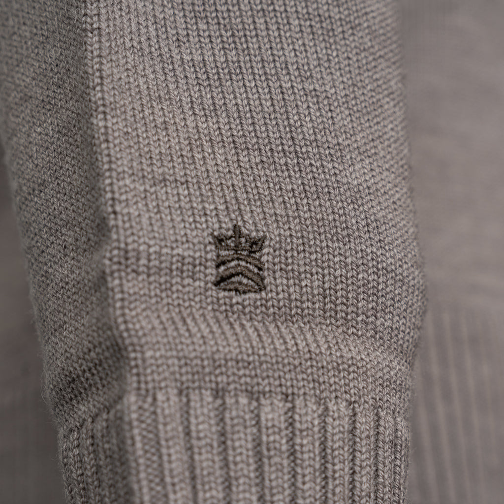 Close-up of a light gray ribbed knit garment, likely a sweater or knit accessory, featuring a small embroidered crown logo in the center. Thomas Maine Knitwear available at Sakks Menswear in Southampton.