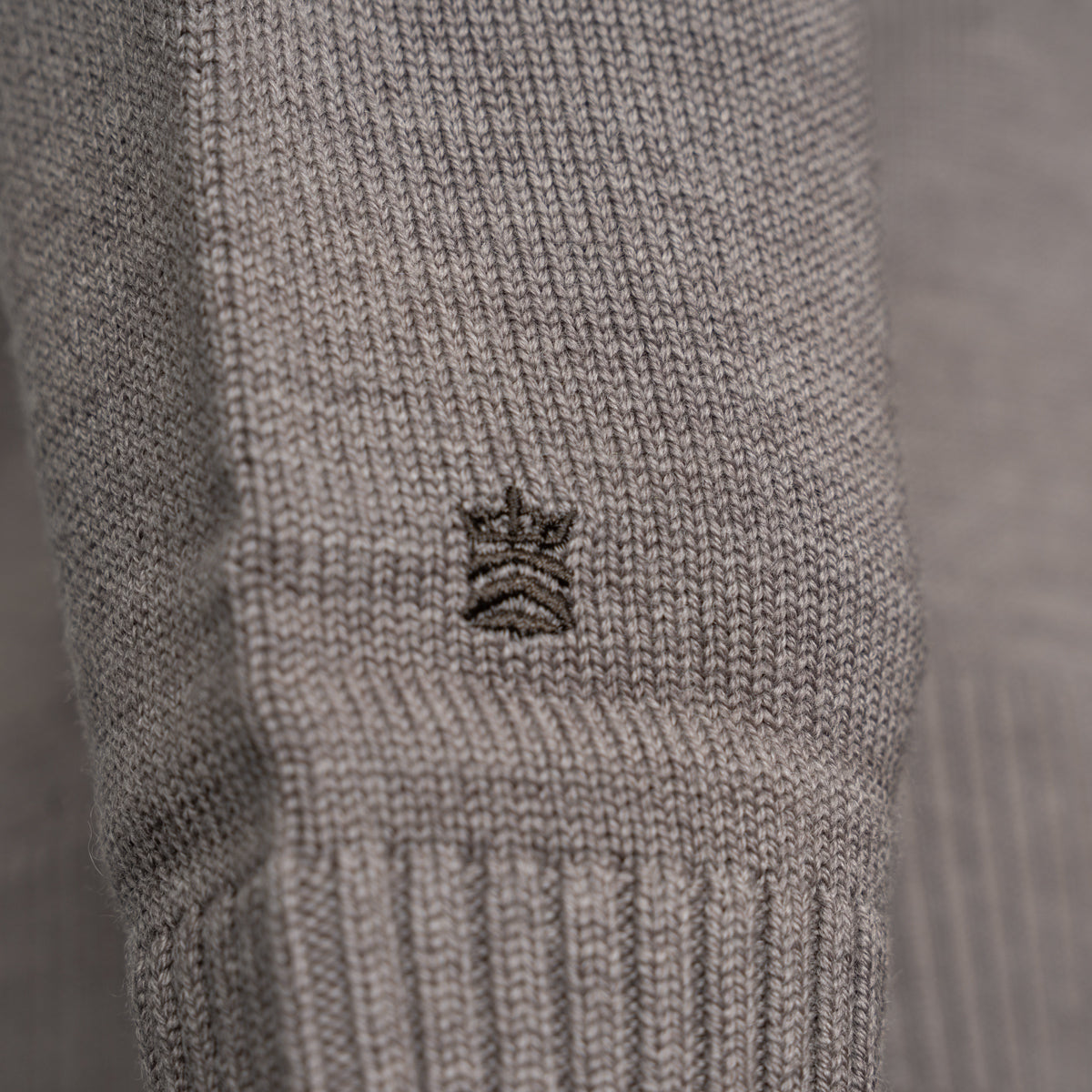 Close-up of a light gray ribbed knit garment, likely a sweater or knit accessory, featuring a small embroidered crown logo in the center. Thomas Maine Knitwear available at Sakks Menswear in Southampton.
