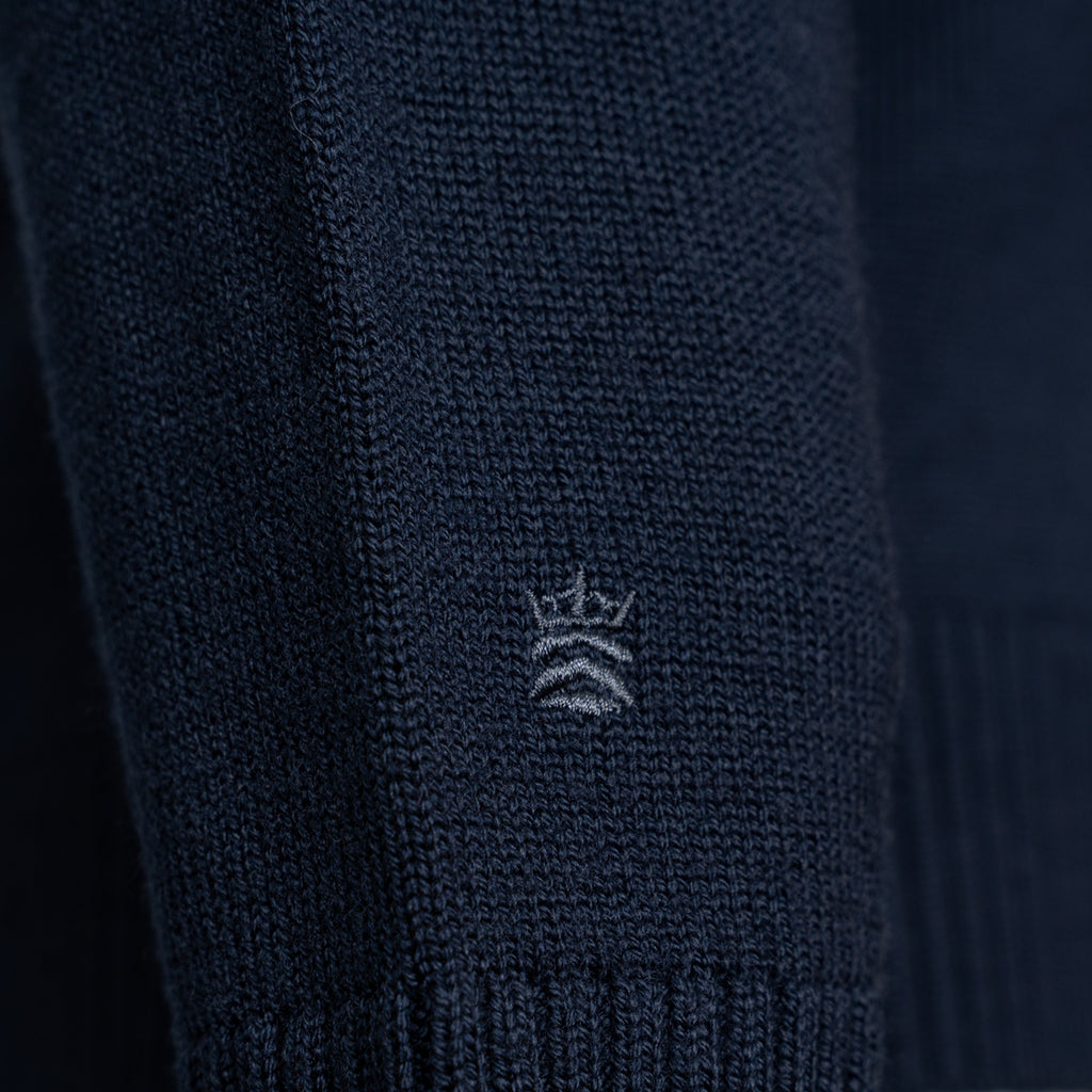 Close-up of a dark navy knit garment, likely a sweater or knit accessory, showing a small white embroidered crown logo on the front. Thomas Maine Knitwear available at Sakks Menswear in Southampton.