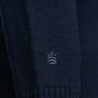 Close-up of a dark navy knit garment, likely a sweater or knit accessory, showing a small white embroidered crown logo on the front. Thomas Maine Knitwear available at Sakks Menswear in Southampton.