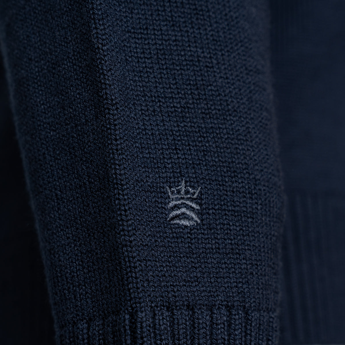 Close-up of a dark navy knit garment, likely a sweater or knit accessory, showing a small white embroidered crown logo on the front. Thomas Maine Knitwear available at Sakks Menswear in Southampton.