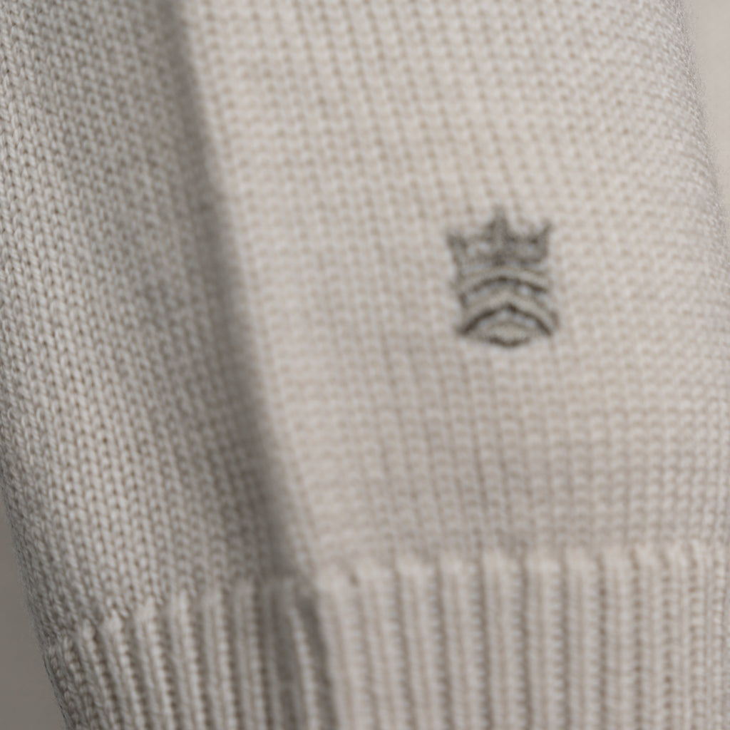 Close-up of a cream-colored knit fabric with a small embroidered gray crown logo on the side. Thomas Maine Knitwear available at Sakks Menswear in Southampton.