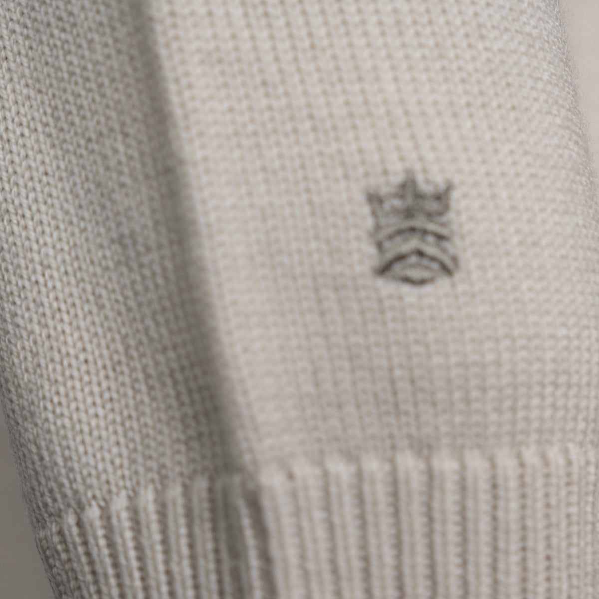 Close-up of a cream-colored knit fabric with a small embroidered gray crown logo on the side. Thomas Maine Knitwear available at Sakks Menswear in Southampton.