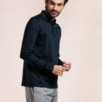 A man wearing a dark navy long-sleeve polo shirt and light gray trousers, standing against a solid light beige background. Thomas Maine Polos available at Sakks Menswear in Southampton.