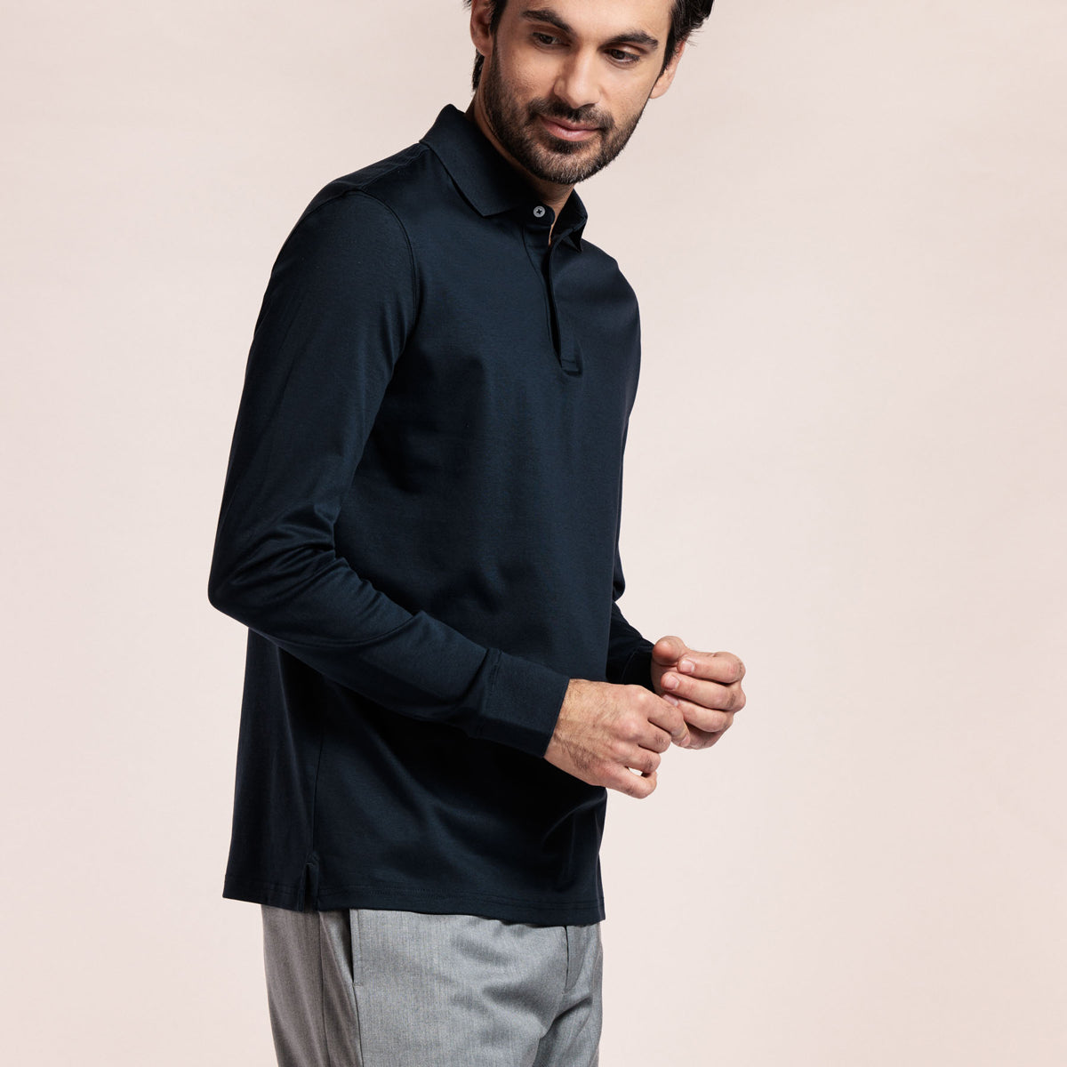 A man wearing a dark navy long-sleeve polo shirt and light gray trousers, standing against a solid light beige background. Thomas Maine Polos available at Sakks Menswear in Southampton.