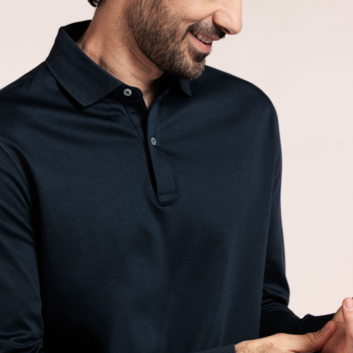 A close-up of a man wearing a dark navy long-sleeve polo shirt against a light beige background. Thomas Maine Polos available at Sakks Menswear in Southampton.