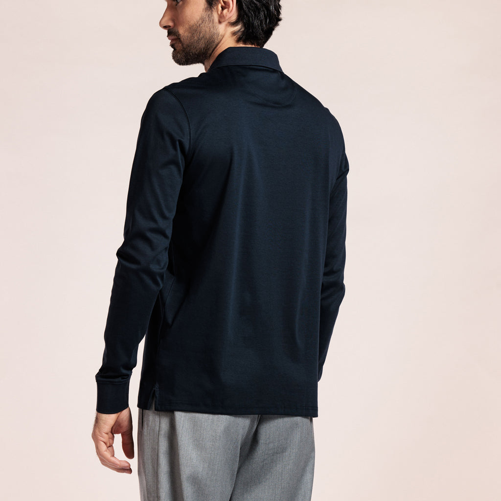 A man wearing a dark navy long-sleeve polo shirt, shown from a rear three-quarter angle against a solid light beige background. Thomas Maine Polos available at Sakks Menswear in Southampton.