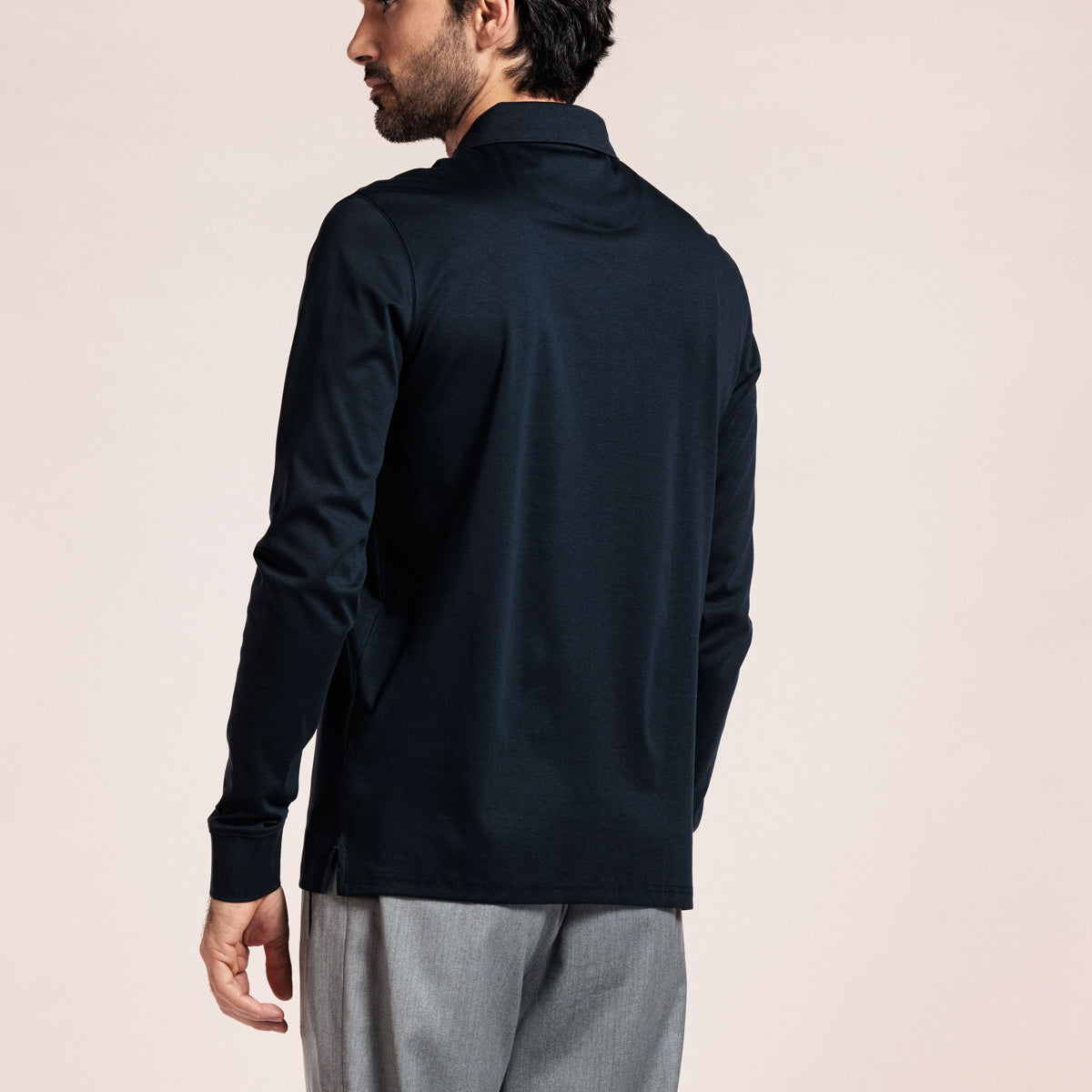 A man wearing a dark navy long-sleeve polo shirt, shown from a rear three-quarter angle against a solid light beige background. Thomas Maine Polos available at Sakks Menswear in Southampton.