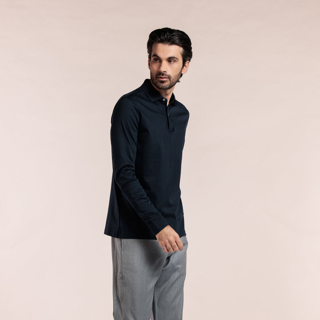 A man wearing a dark navy long-sleeve polo shirt and light gray trousers, standing against a solid pale pink studio background. Thomas Maine Polos available at Sakks Menswear in Southampton.