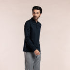 A man wearing a dark navy long-sleeve polo shirt and light gray trousers, standing against a solid pale pink studio background. Thomas Maine Polos available at Sakks Menswear in Southampton.