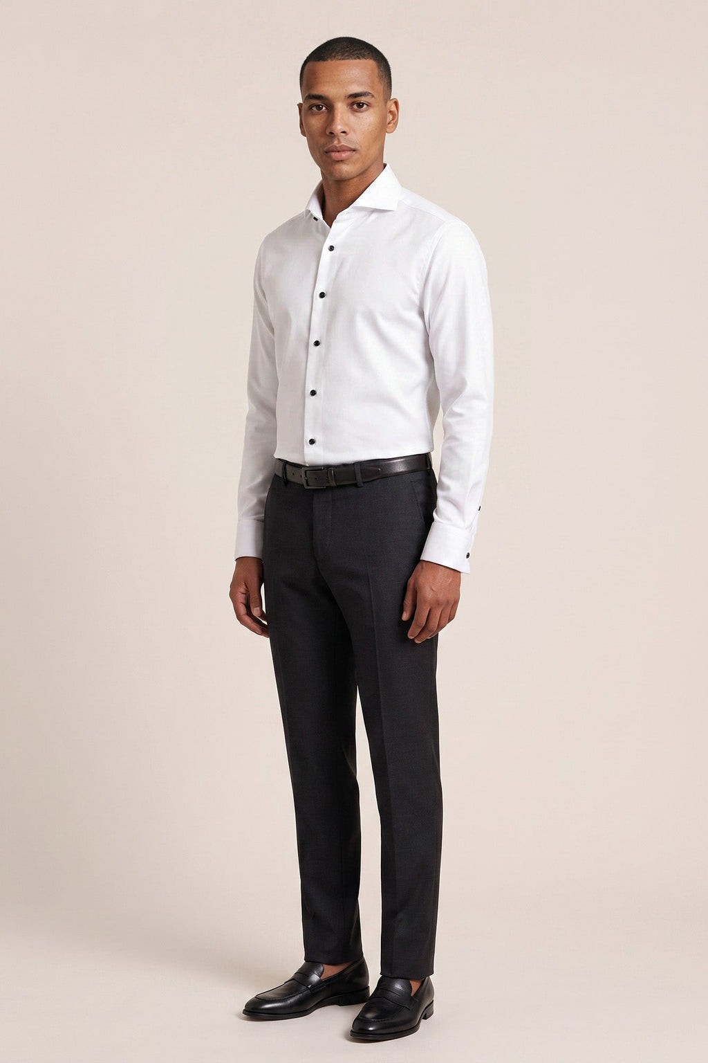 A man modeling a white long-sleeve button-down dress shirt with black trousers, a black belt, and black dress shoes, standing against a solid light beige studio background. Thomas Maine shirts available at Sakks Menswear in Southampton.