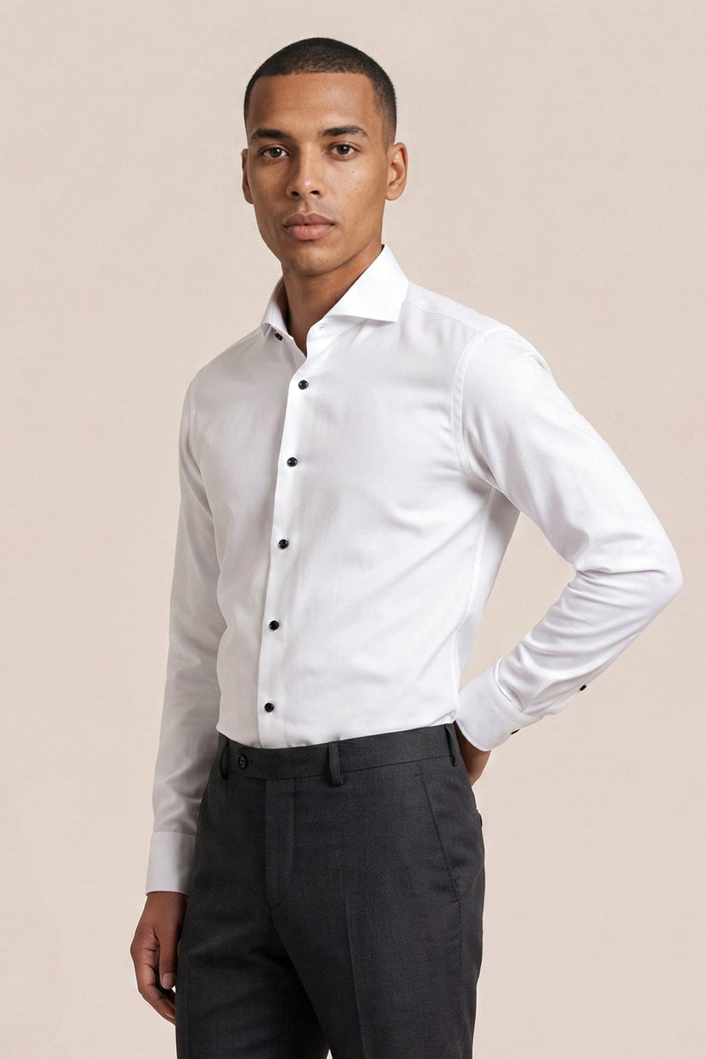 A man wearing a white long-sleeve button-down dress shirt and dark gray trousers, standing against a solid light beige background. Thomas Maine shirts available at Sakks Menswear in Southampton.
