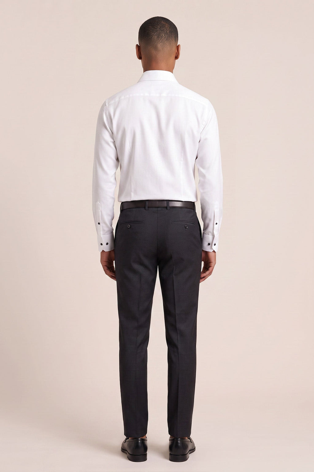 A man viewed from the back wearing a white long-sleeve dress shirt, dark charcoal trousers, a black belt, and black dress shoes, against a solid light beige studio background. Thomas Maine shirts available at Sakks Menswear in Southampton.