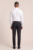 A man viewed from the back wearing a white long-sleeve dress shirt, dark charcoal trousers, a black belt, and black dress shoes, against a solid light beige studio background. Thomas Maine shirts available at Sakks Menswear in Southampton.