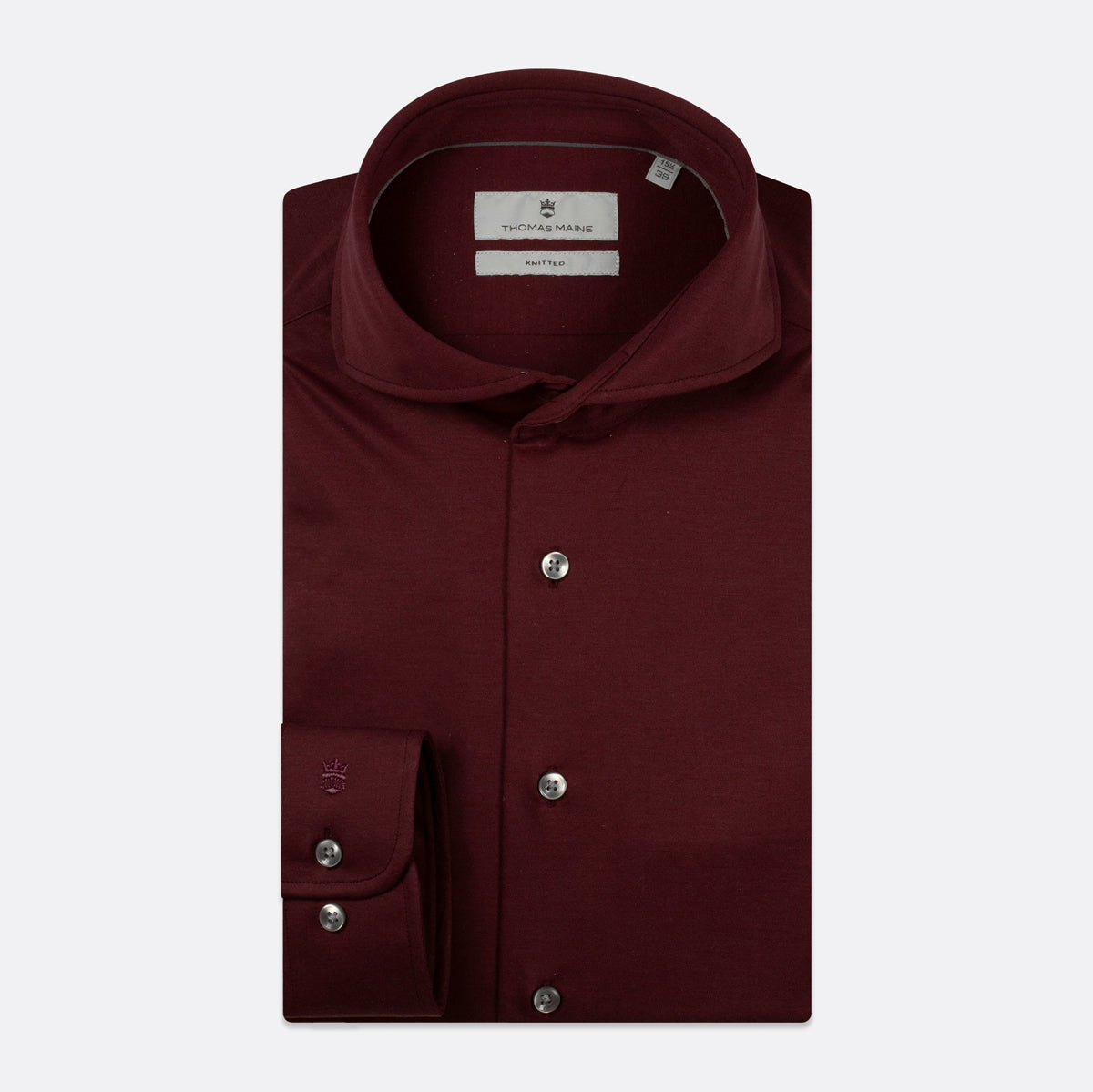 A folded burgundy long-sleeve dress shirt by Thomas Maine, featuring a pointed collar, silver buttons, and a single cuff with two buttons, displayed on a light gray background. Thomas Maine Shirts available at Sakks Menswear in Southampton.