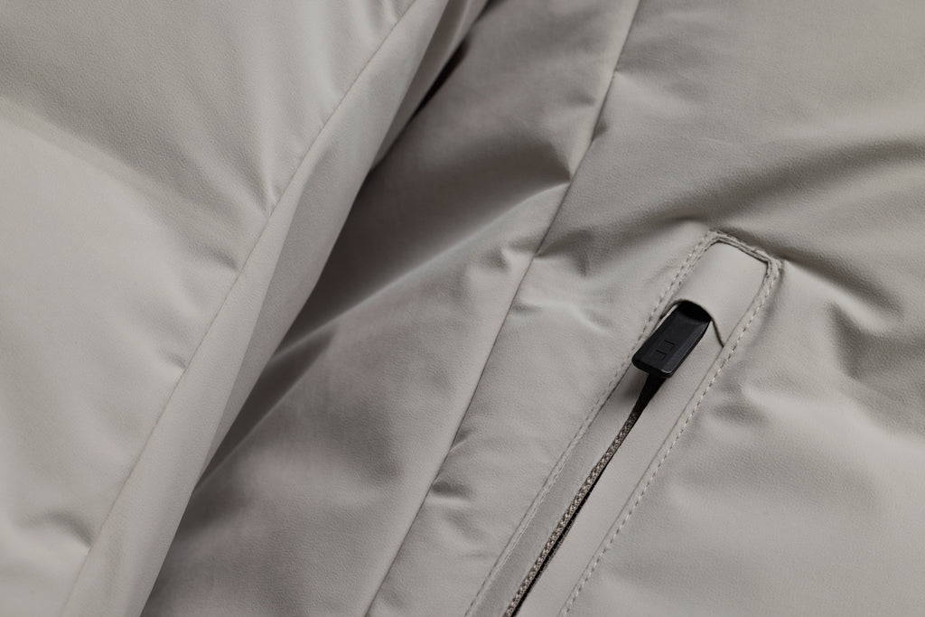 Close-up of a light gray puffer jacket showing a zippered pocket with a black zipper pull. UBR Jackets available at Sakks Menswear in Southampton.