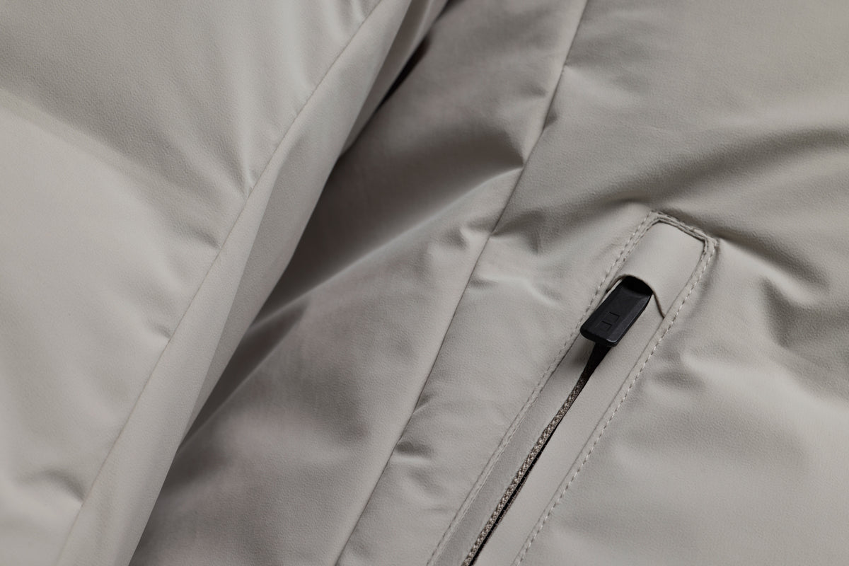 Close-up of a light gray puffer jacket showing a zippered pocket with a black zipper pull. UBR Jackets available at Sakks Menswear in Southampton.
