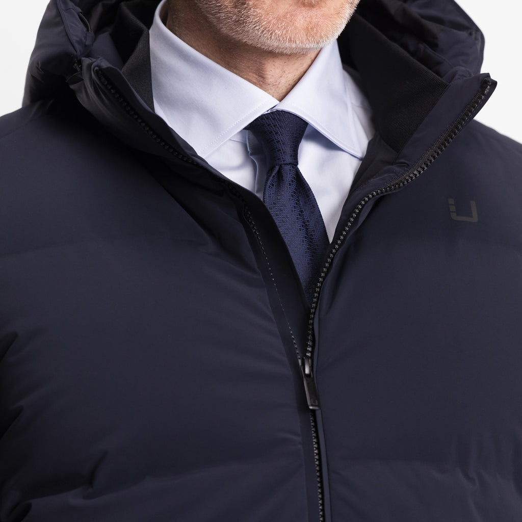 Model wearing the UBR Bolt down Jacket in Navy. The image is a close up of the zip on the puffer coat. The model is wearing the down jacket with a white dress shirt and navy blue tie. The model is stood in front of a light grey background. UBR Jackets available at Sakks Menswear in Southampton.