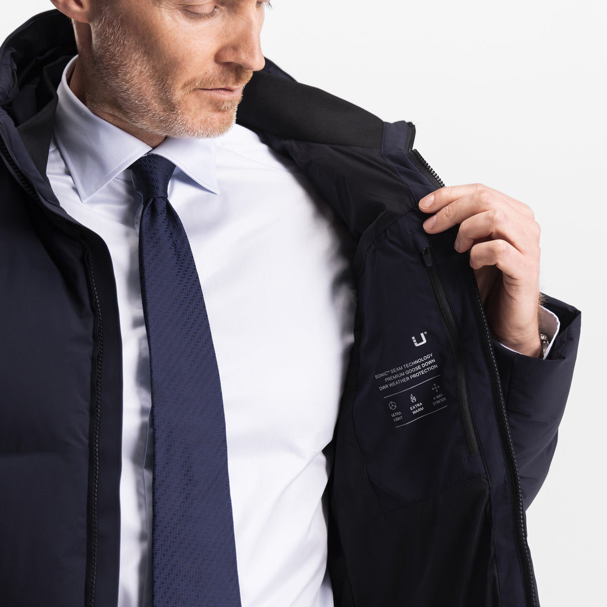 Model wearing the UBR Bolt down Jacket in Navy. Model is wearing the puffer coat with a white dress shirt and navy blue tie. The model is stood in front of a light grey background. UBR Jackets available at Sakks Menswear in Southampton.