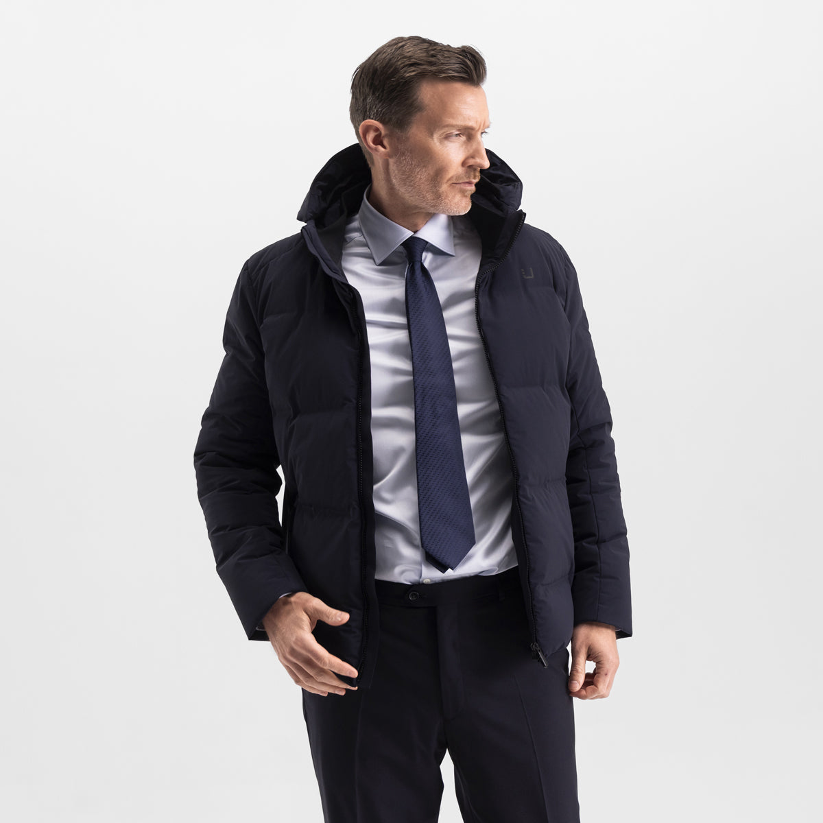 Model wearing the UBR Bolt down Jacket in Navy. Model is wearing the puffer coat with a pair of smart navy trousers. The model is stood in front of a light grey background. UBR Jackets available at Sakks Menswear in Southampton.