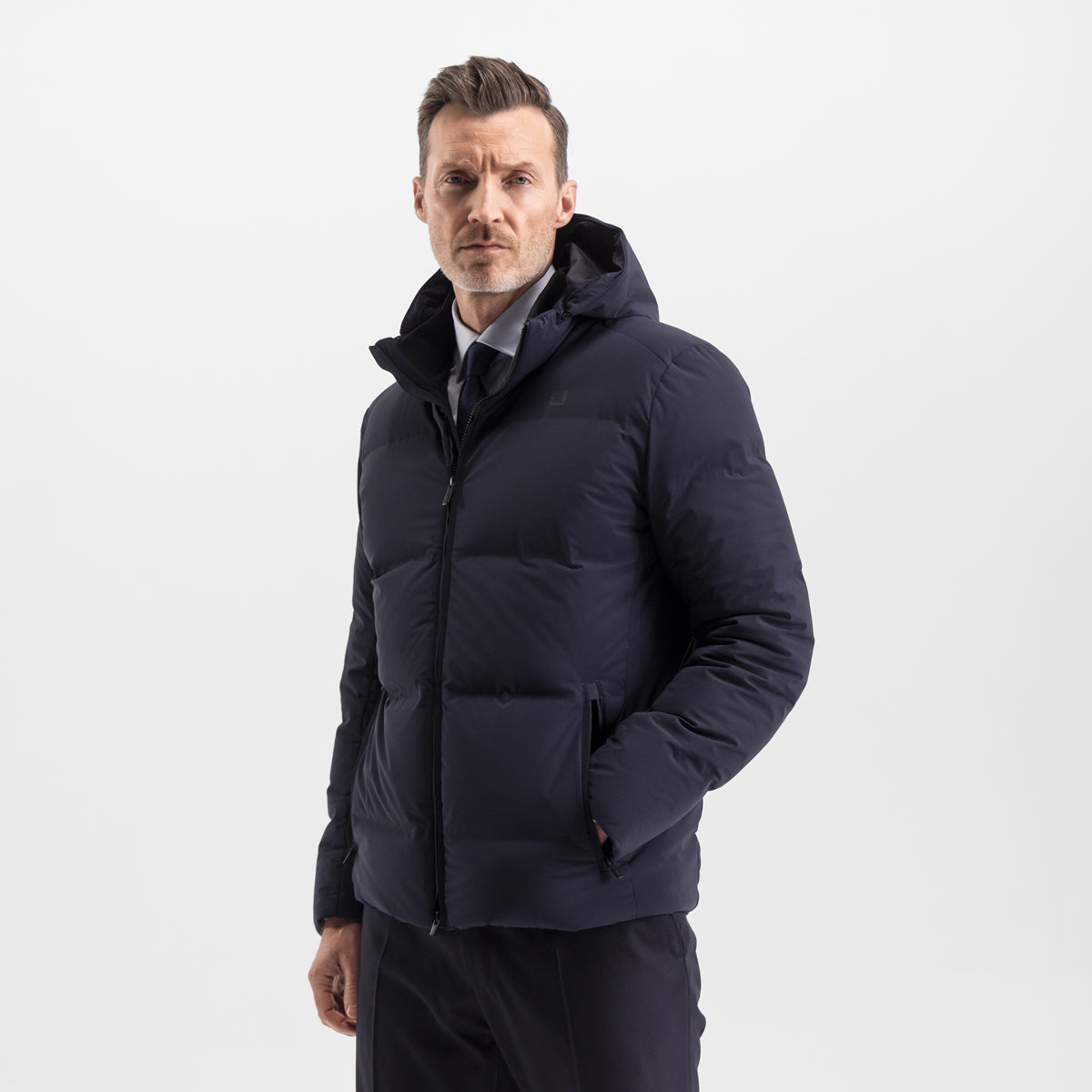 Model wearing the UBR Bolt down Jacket in Navy. Model is wearing the puffer coat with a pair of smart navy trousers. The model is stood in front of a light grey background. UBR Jackets available at Sakks Menswear in Southampton.