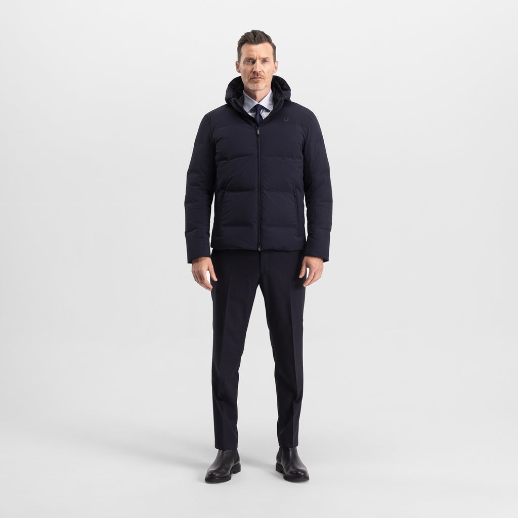 Model wearing the UBR Bolt down Jacket in Navy. Model is wearing the puffer coat with a pair of smart navy trousers and black chelsea boots. The model is stood in front of a light grey background. UBR Jackets available at Sakks Menswear in Southampton.