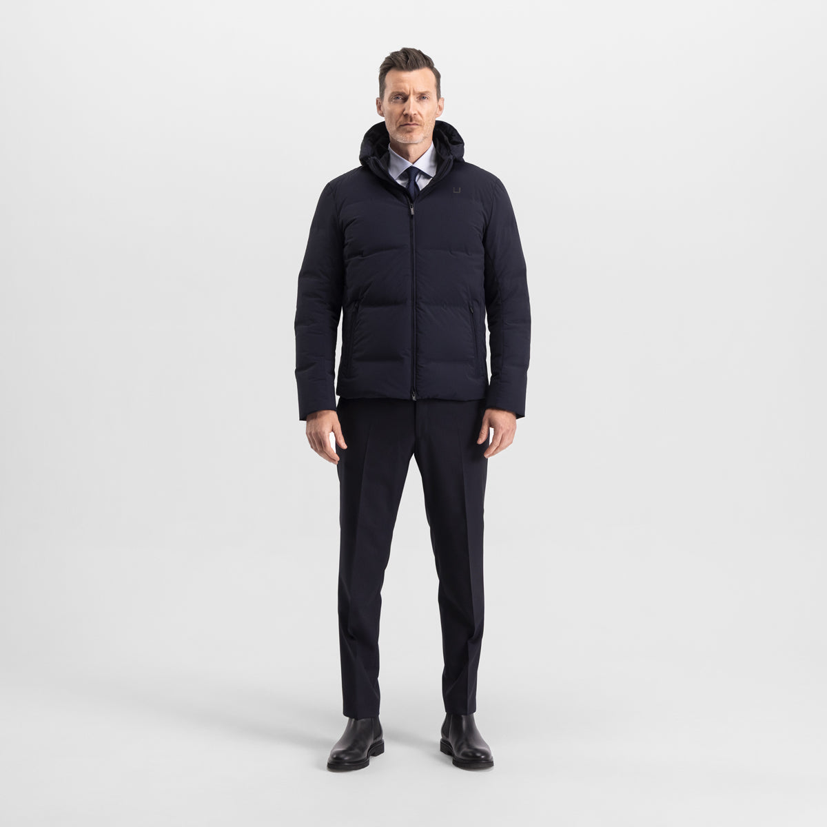 Model wearing the UBR Bolt down Jacket in Navy. Model is wearing the puffer coat with a pair of smart navy trousers and black chelsea boots. The model is stood in front of a light grey background. UBR Jackets available at Sakks Menswear in Southampton.