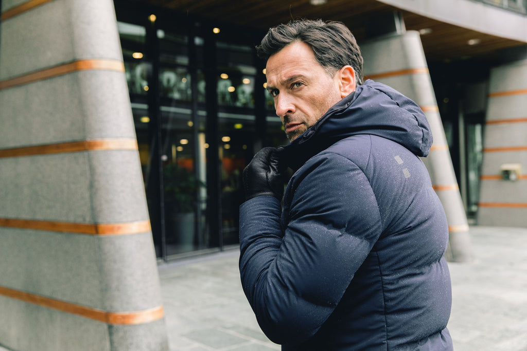 Image of a man wearing a navy blue puffer coat. The model is pictured zipping up his navy UBR Bolt down Jacket. The model is pictured in front of a stone pathway in front of grey buildings. UBR Jackets available at Sakks Menswear in Southampton.