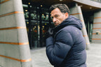 Image of a man wearing a navy blue puffer coat. The model is pictured zipping up his navy UBR Bolt down Jacket. The model is pictured in front of a stone pathway in front of grey buildings. UBR Jackets available at Sakks Menswear in Southampton.