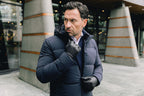 Image of a man wearing a navy blue puffer coat. The model is pictured zipping up his navy UBR Bolt down Jacket. The model is pictured in front of a stone pathway in front of grey buildings. UBR Jackets available at Sakks Menswear in Southampton.