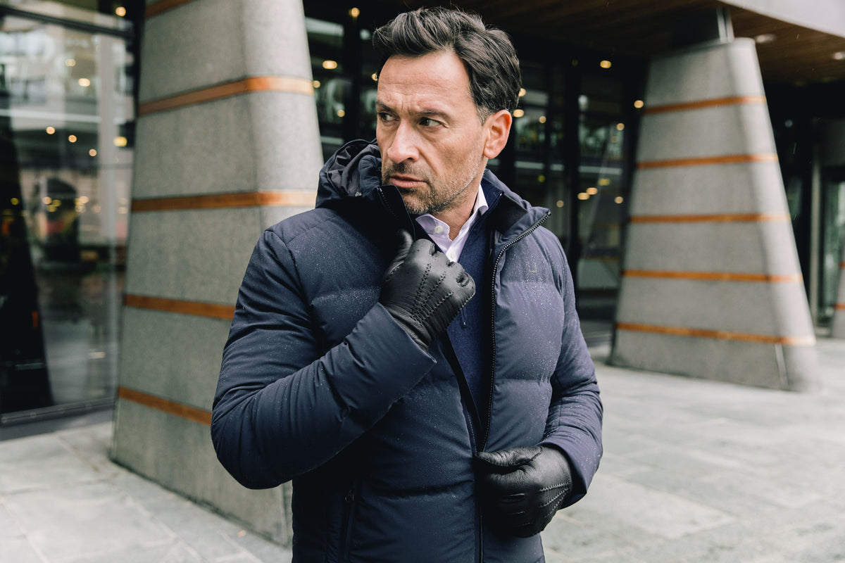 Image of a man wearing a navy blue puffer coat. The model is pictured zipping up his navy UBR Bolt down Jacket. The model is pictured in front of a stone pathway in front of grey buildings. UBR Jackets available at Sakks Menswear in Southampton.