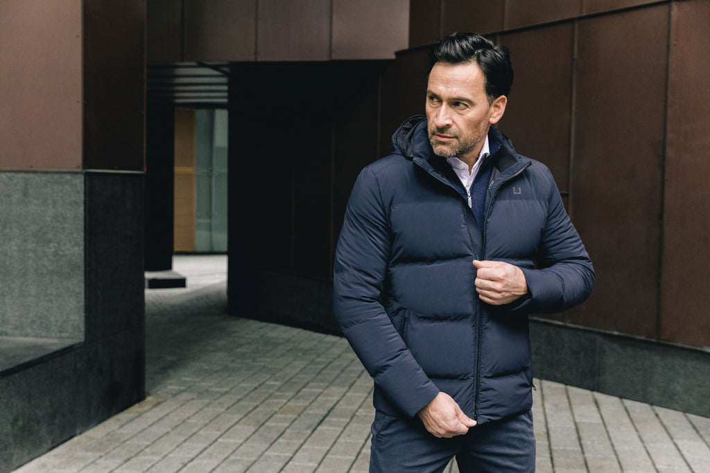 Image of a man wearing a navy blue puffer coat. The model is pictured zipping up his navy UBR Bolt down Jacket. The model is pictured in front of a stone pathway in front of brown buildings. UBR Jackets available at Sakks Menswear in Southampton.