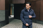 Image of a man wearing a navy blue puffer coat. The model is pictured zipping up his navy UBR Bolt down Jacket. The model is pictured in front of a stone pathway in front of brown buildings. UBR Jackets available at Sakks Menswear in Southampton.