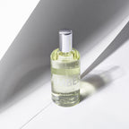 A clear glass perfume bottle filled with pale yellow liquid, featuring a reflective silver cap and a semi-transparent white label that reads 'AMBER'. The bottle is positioned on a white surface under sharp, dramatic diagonal shadows. Laboratory Perfumes Amber available at Sakks Menswear in Southampton.