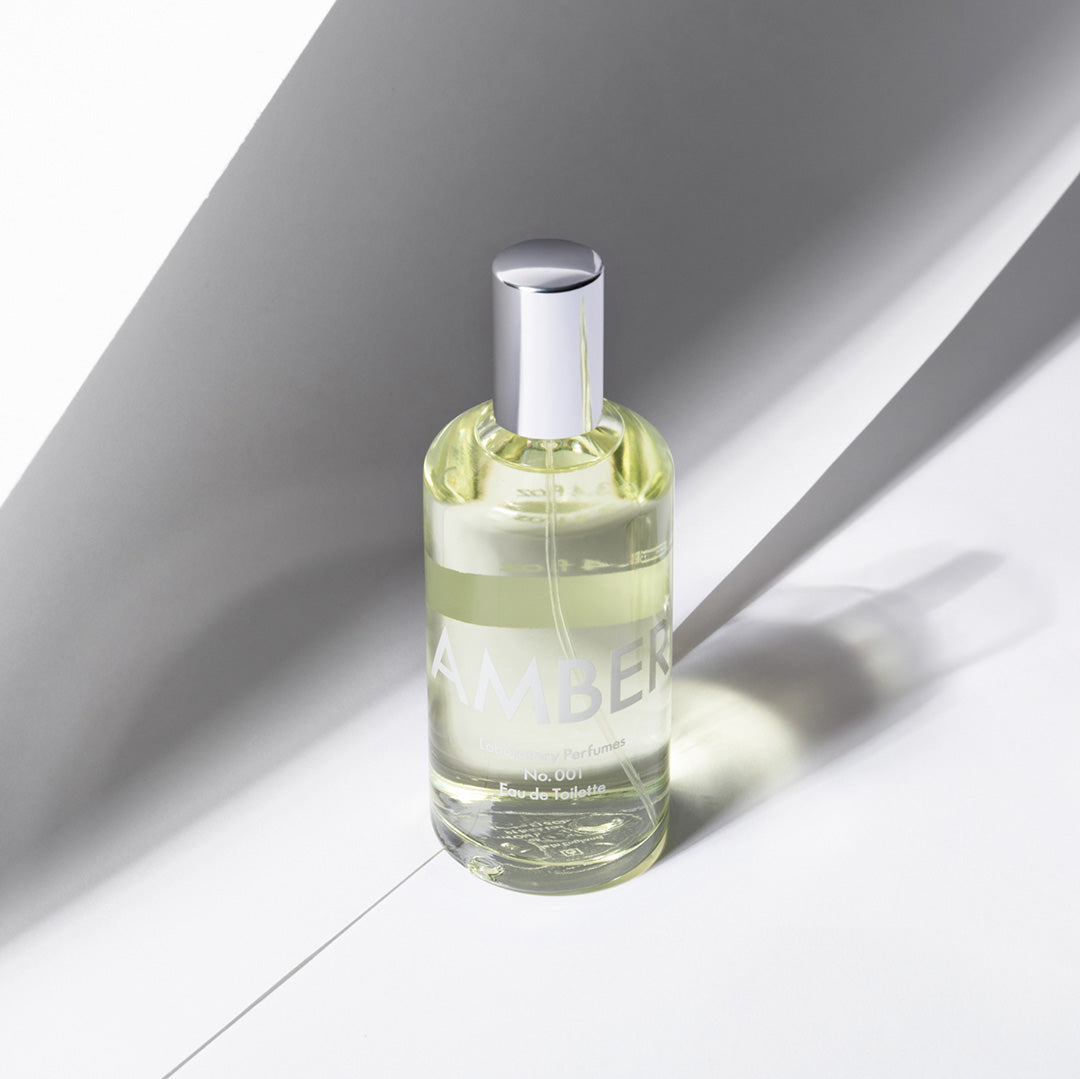 A clear glass perfume bottle filled with pale yellow liquid, featuring a reflective silver cap and a semi-transparent white label that reads 'AMBER'. The bottle is positioned on a white surface under sharp, dramatic diagonal shadows. Laboratory Perfumes Amber available at Sakks Menswear in Southampton.