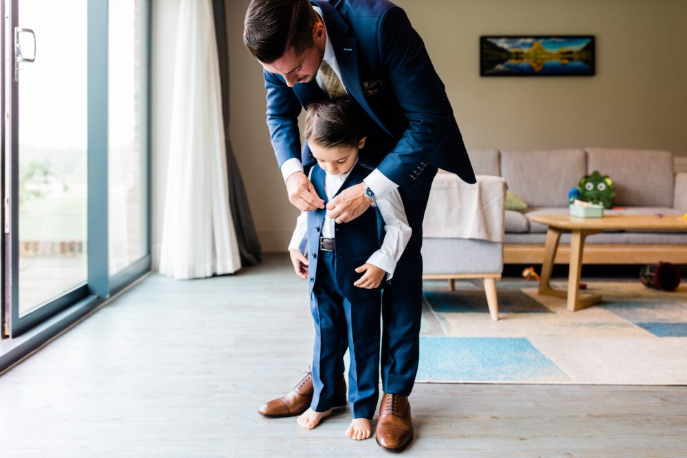 A man in a navy blue suit helps a young boy put on a matching navy blue suit and tie in a modern living room setting. Sakks Menswear Made-To-Measure Waistcoat in Southampton.