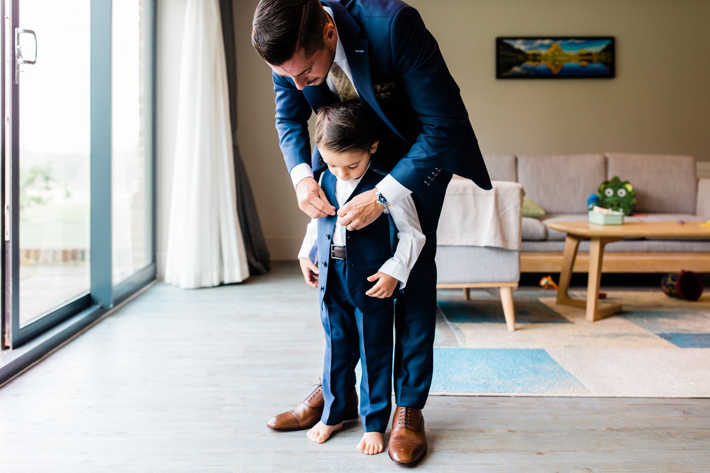 A man in a navy blue suit helps a young boy put on a matching navy blue suit and tie in a modern living room setting. Sakks Menswear Made-To-Measure in Southampton.