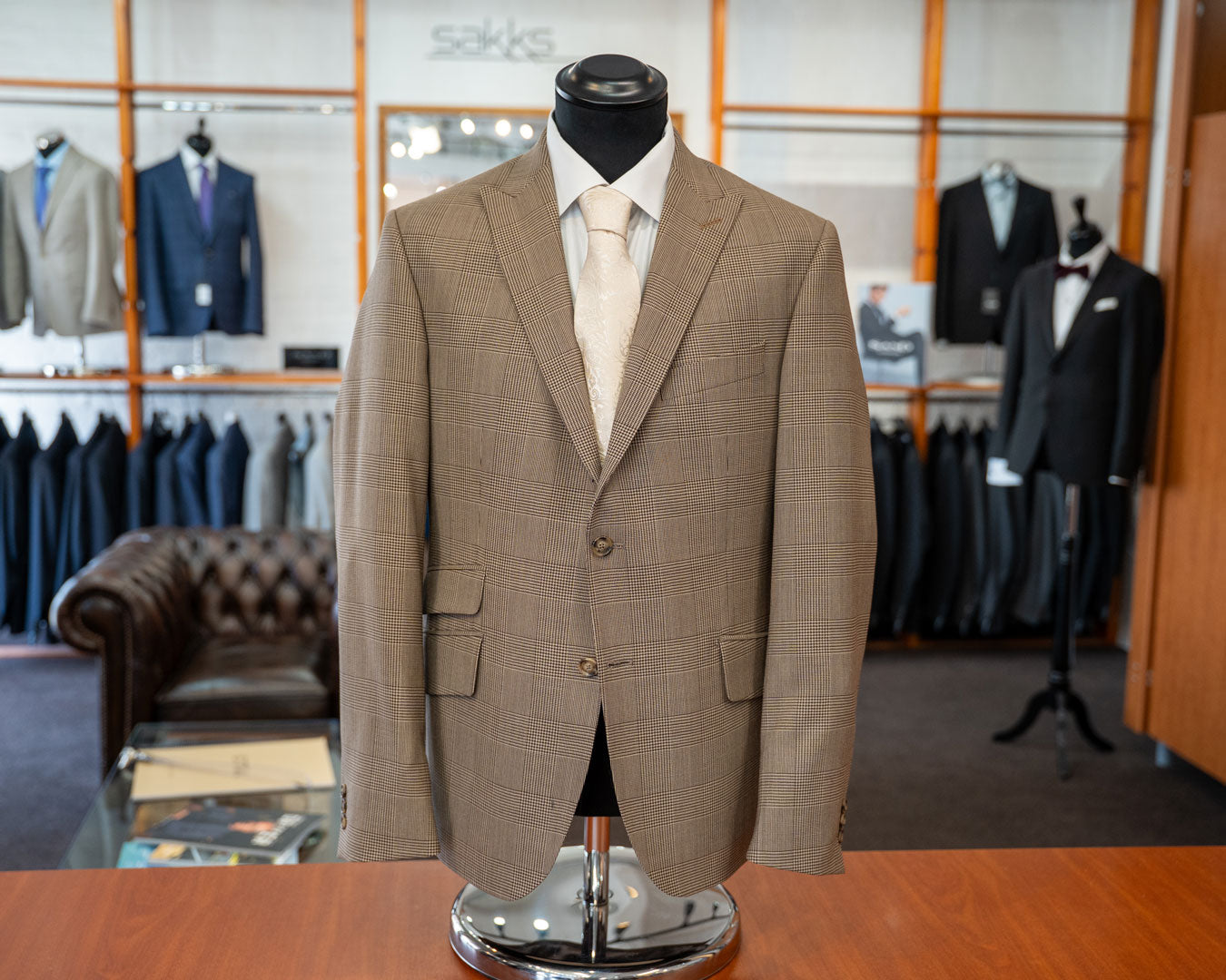 A men's tailored suit jacket displayed on a black mannequin torso in a clothing boutique. The jacket is light tan with a subtle windowpane plaid pattern, paired with a white dress shirt and cream tie. Sakks Menswear Made-To-Measure Service in Southampton.