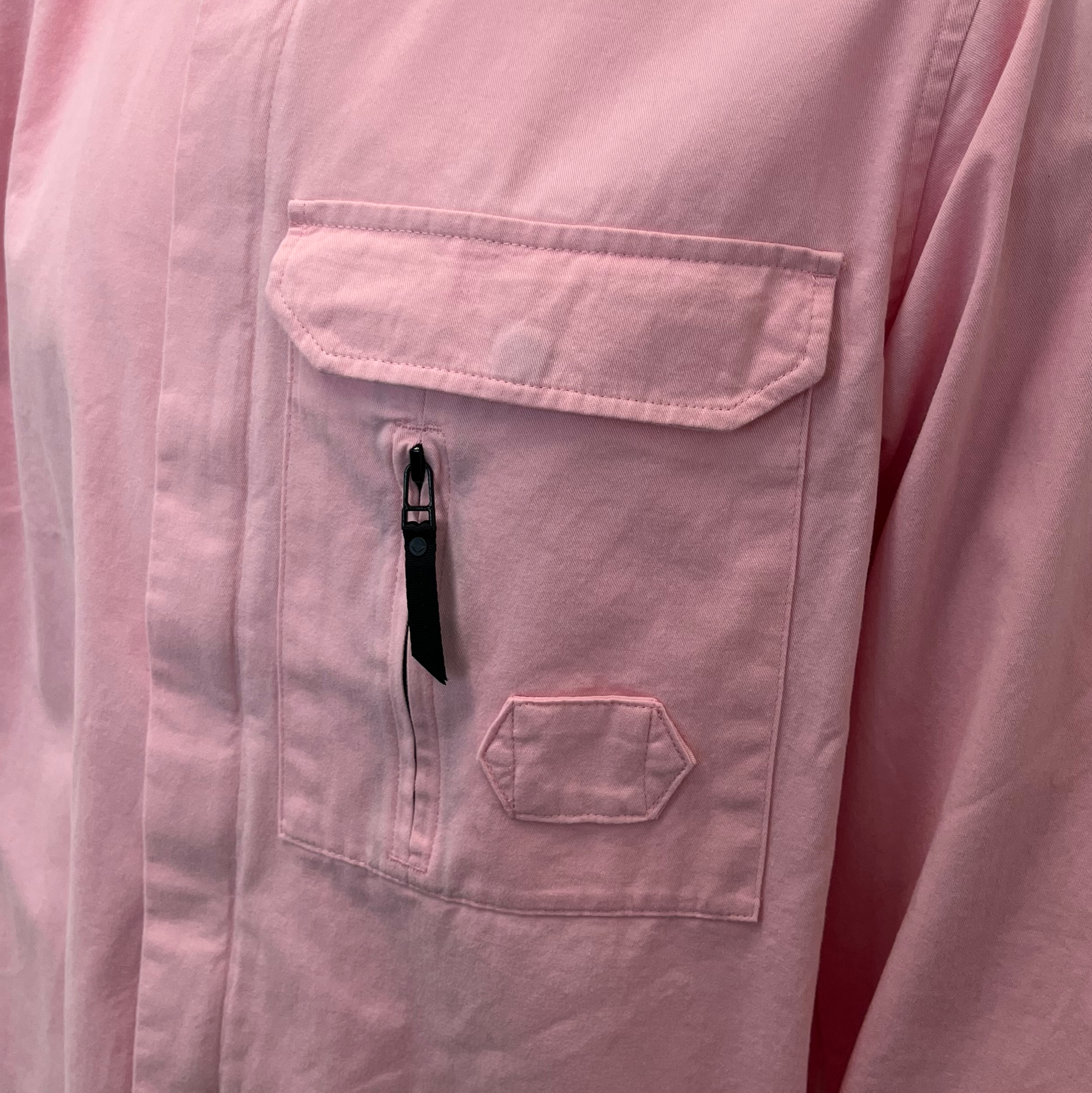 Close-up of a light pink long-sleeve shirt or jacket showing a chest pocket with a flap, a vertical zippered pocket with a black zipper pull, and a small hexagonal stitched pocket below. MA.Strum Shirt Jacket available at Sakks Menswear in Southampton.
