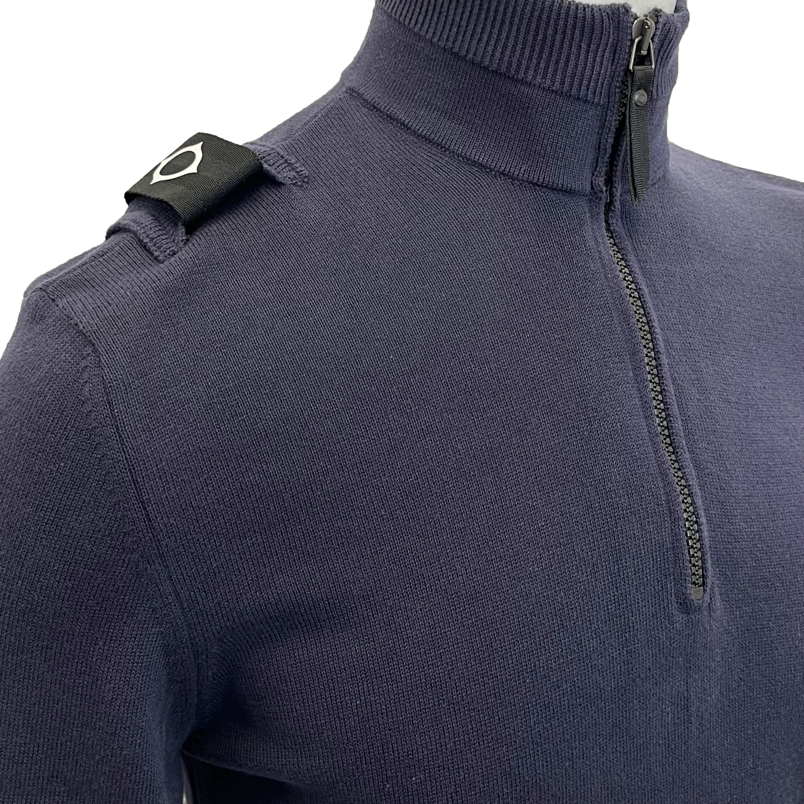Close-up of a navy blue ribbed knit quarter-zip sweater with a black shoulder tab featuring a white geometric logo. MA.Strum Knitwear available at Sakks Menswear in Southampton.