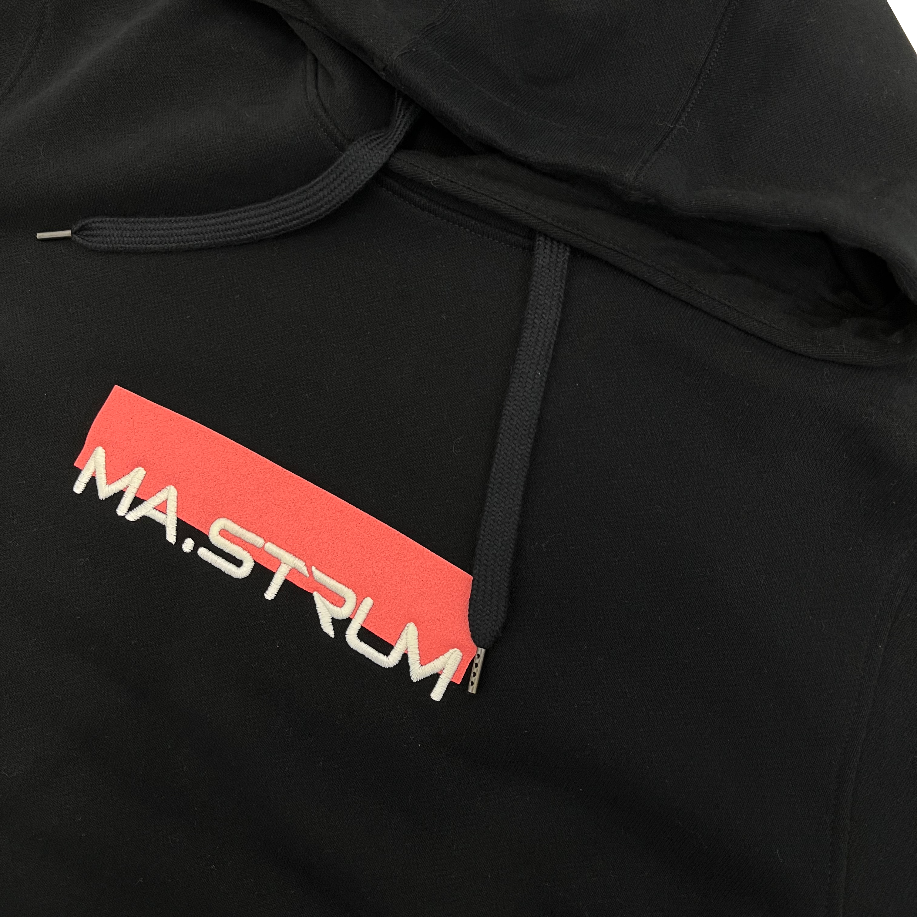 Close-up of a black hoodie featuring a red rectangular chest graphic with the text 'MA.STRUM' in white. MA.Strum Hoodie available at Sakks Menswear in Southampton.