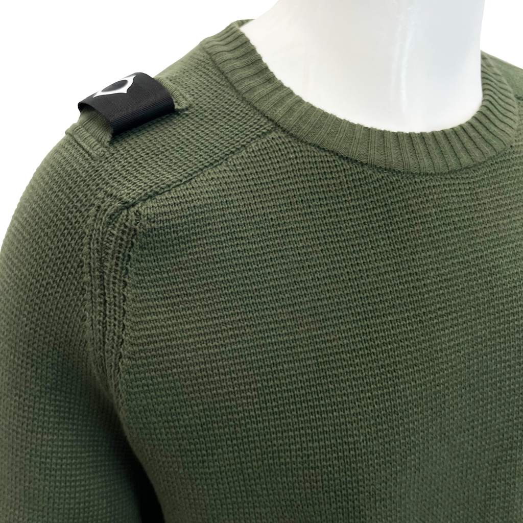 Close-up of an olive green ribbed knit sweater on a white mannequin torso, showing a black shoulder patch with a white stylised logo. MA.Strum Jumper available at Sakks Menswear in Southampton.