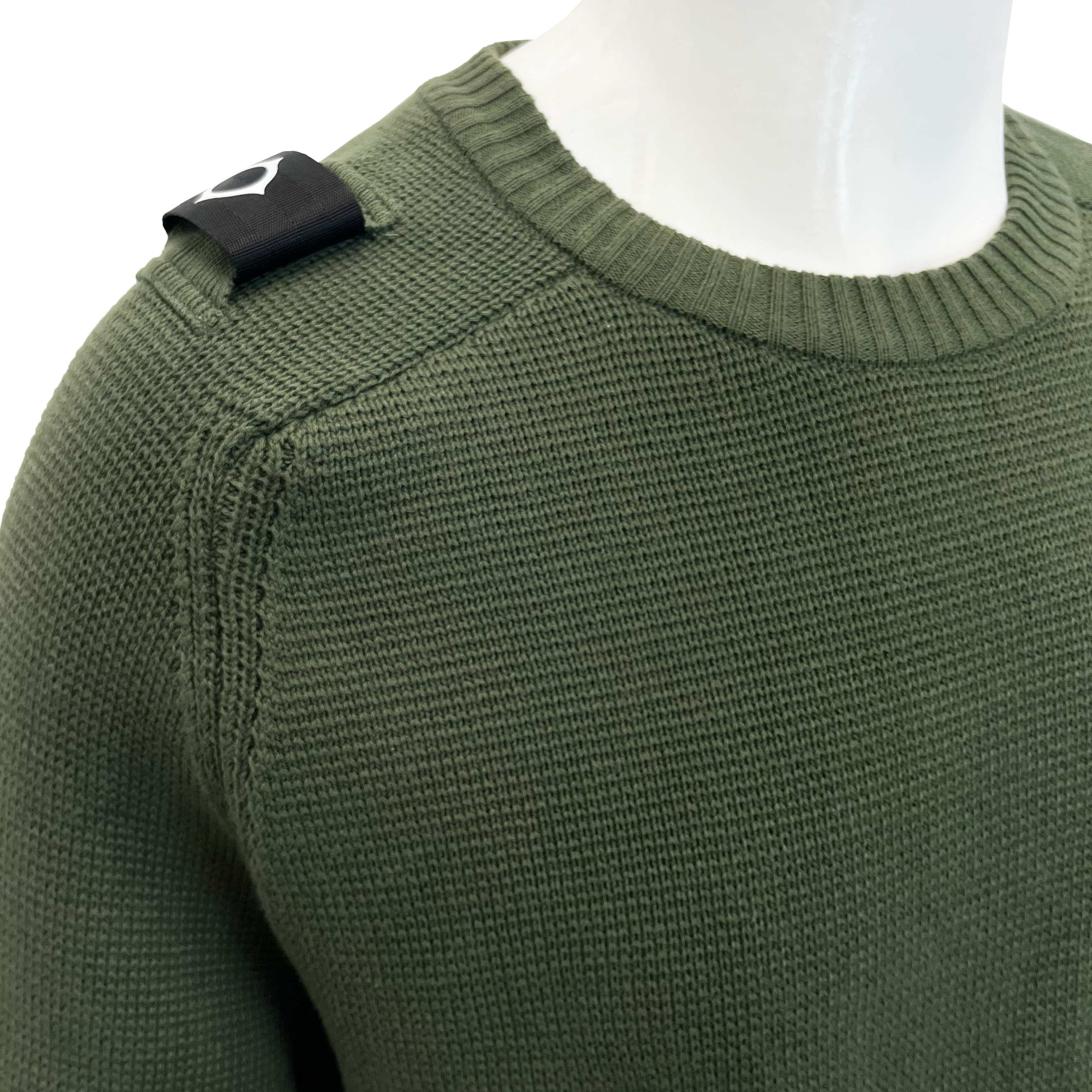 Close-up of an olive green ribbed knit sweater on a white mannequin torso, showing a black shoulder patch with a white stylised logo. MA.Strum Jumper available at Sakks Menswear in Southampton.