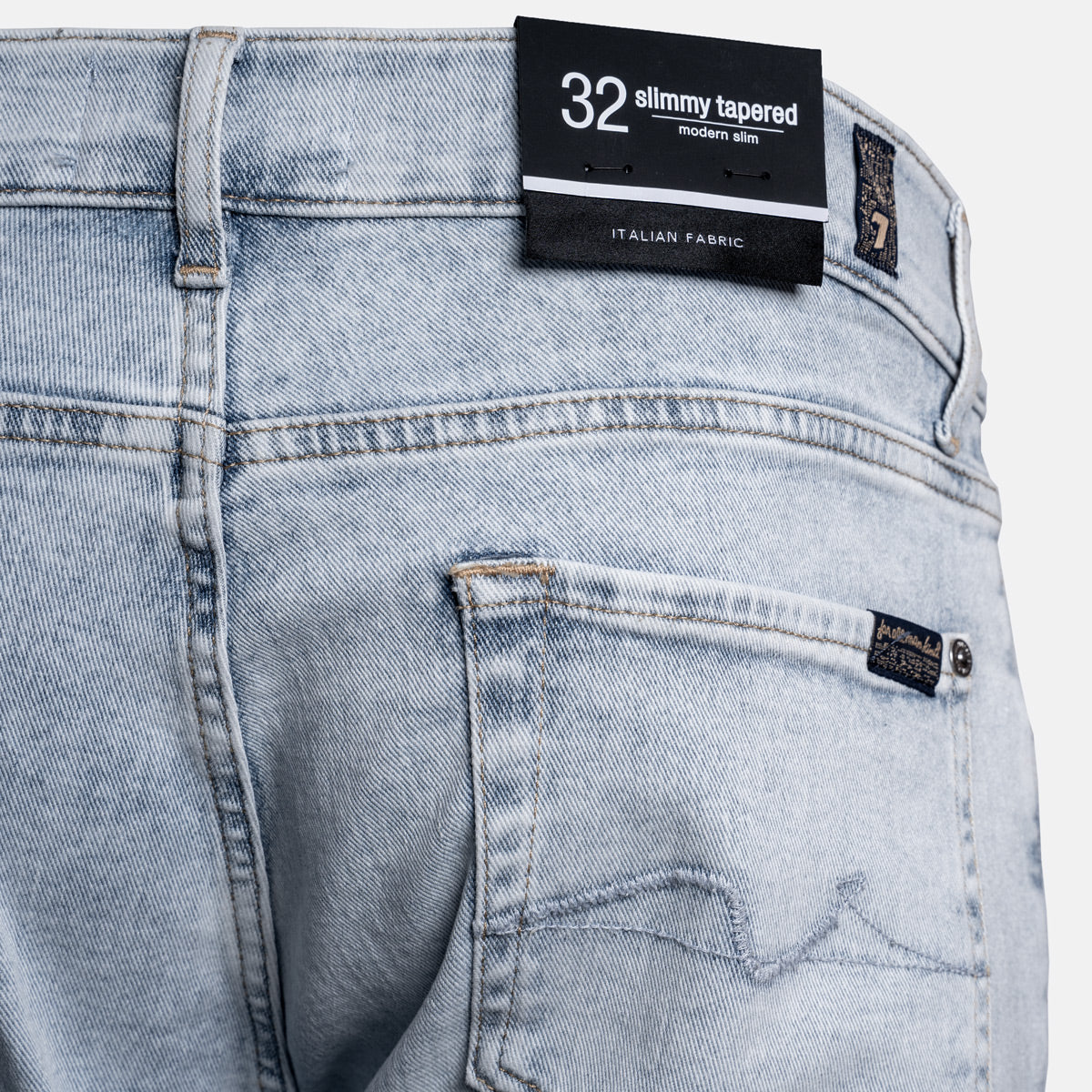 Close-up of the back waistband and pocket area of light-wash denim jeans, showing a black size label reading 32 slimmy tapered and a small brand patch on the pocket. 7 For All Mankind Jeans available at Sakks Menswear in Southampton.