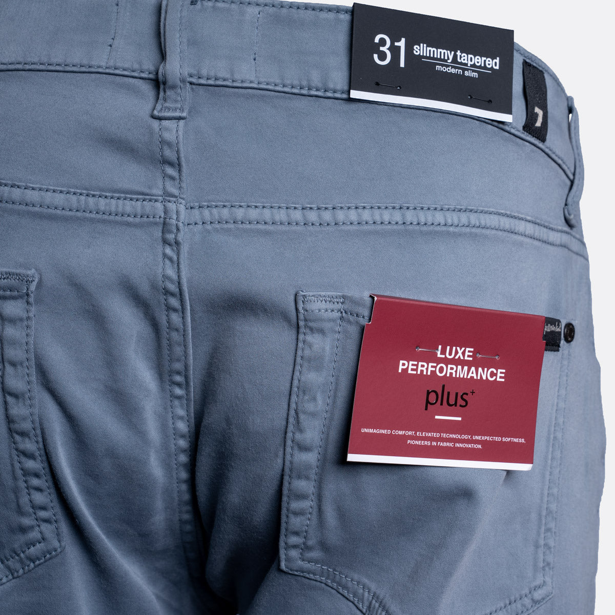 Close-up of the back waistband and pocket area of slate blue tapered pants, showing a black size label reading '31 slimmy tapered' and a red fabric tag reading 'LUXE PERFORMANCE plus'. 7 For All Mankind Chinos available at Sakks Menswear in Southampton.